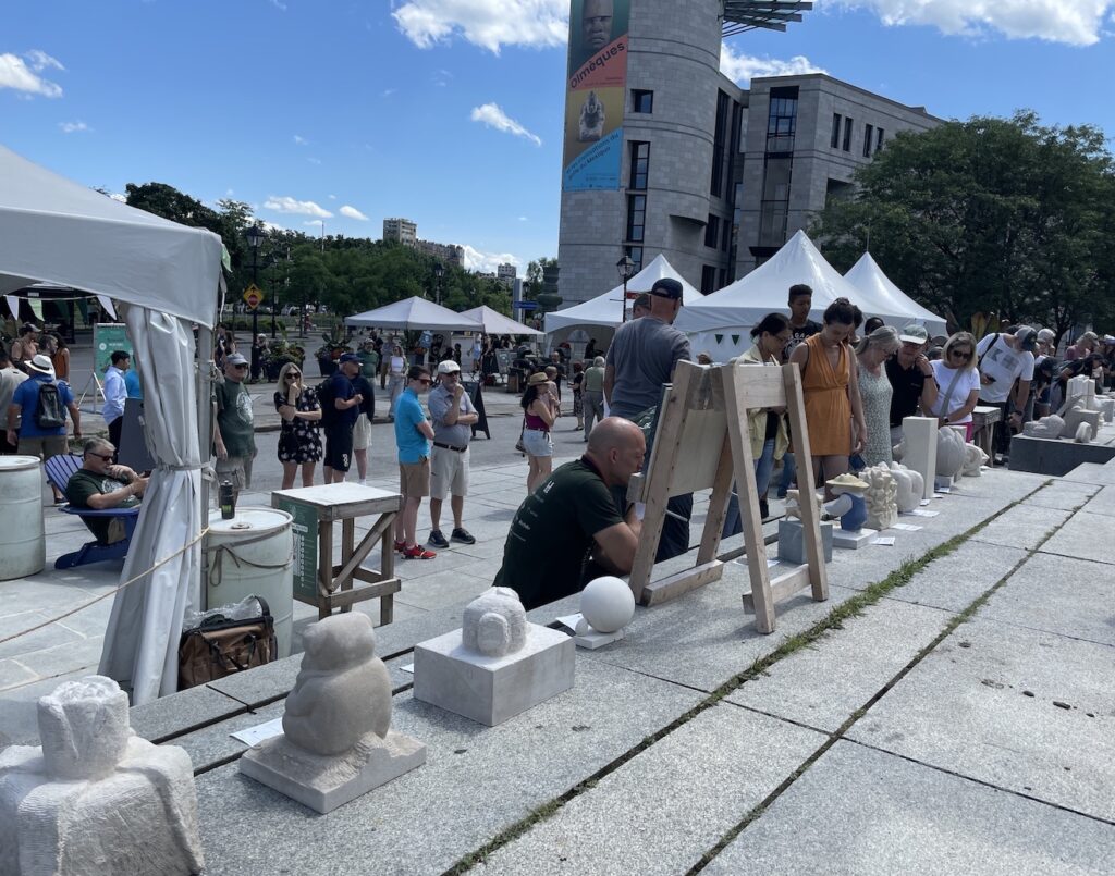 Stone sculptures on display at an outdoor auction in a city, surrounded by people
