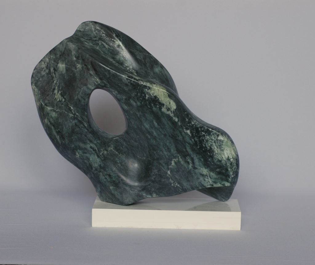 Evergreen_1 Abstract dark green marbled stone sculpture with a wavy, twisting silhouette and an oval opening, on a light beige stone base.
