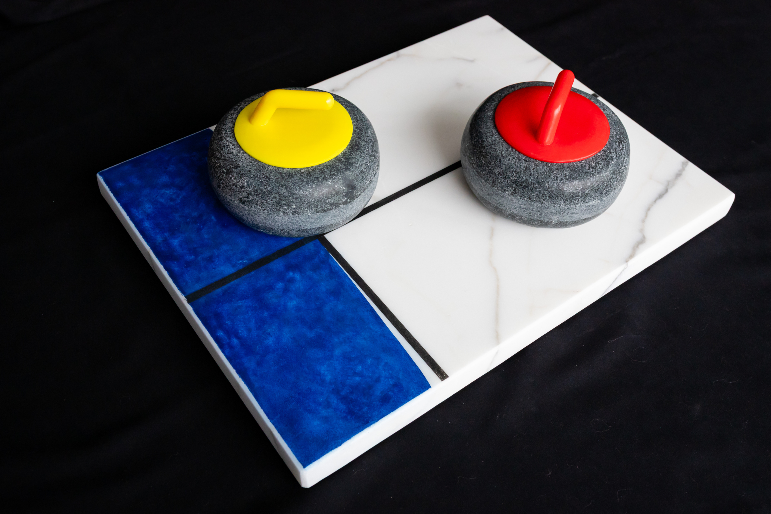 Two replica curling stones with yellow and red handles resting on a white marble base with a part painted blue.