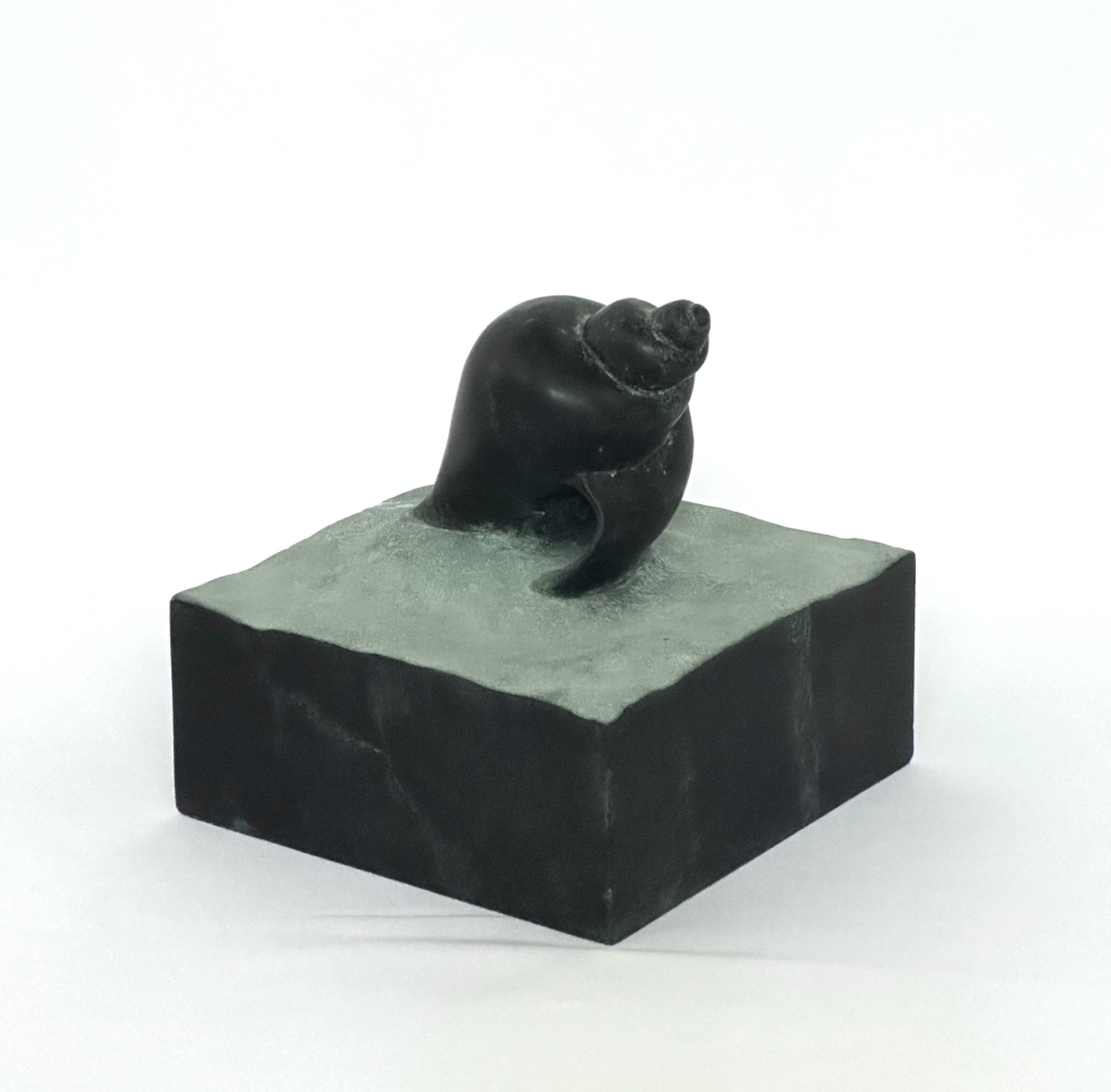 Quarry_1.jpg Small dark stone sculpture of a conch shell, set on a grey stone base.